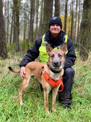 Man kneeling in hi vis vest with a K9 sniffer dog in front looking at camera